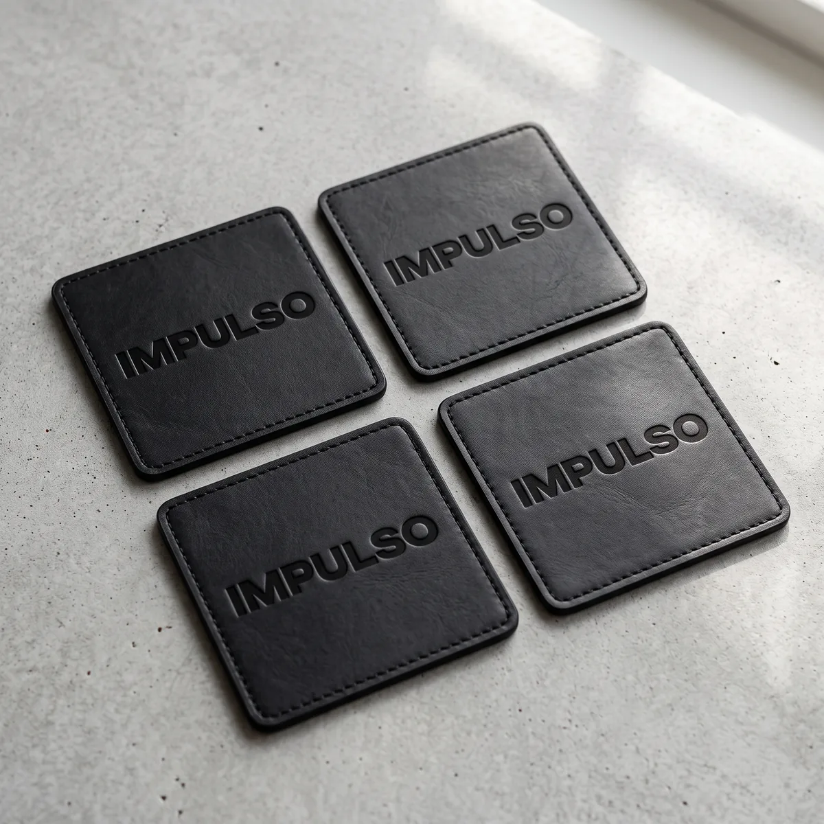 Four square black leather coasters with the IMPULSO wordmark debossed, laid out on concrete