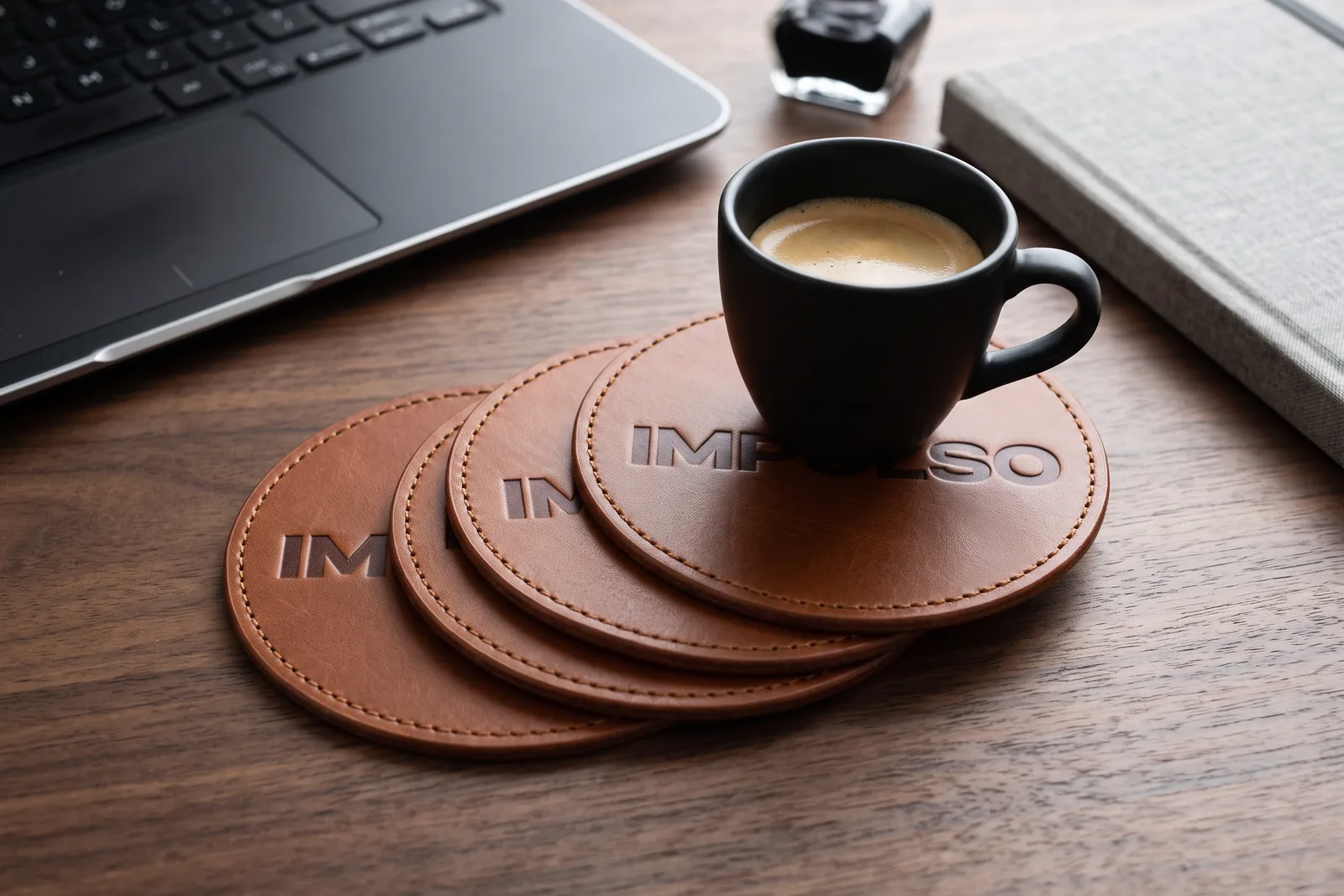 Cognac leather coasters with the IMPULSO wordmark debossed into the leather, under an espresso cup on a walnut desk