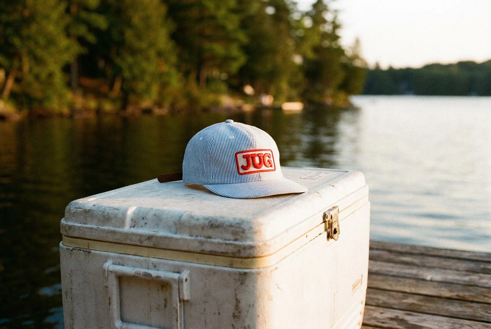 A JUG striped-brim hat resting on a vintage cooler on a Muskoka dock at sunset