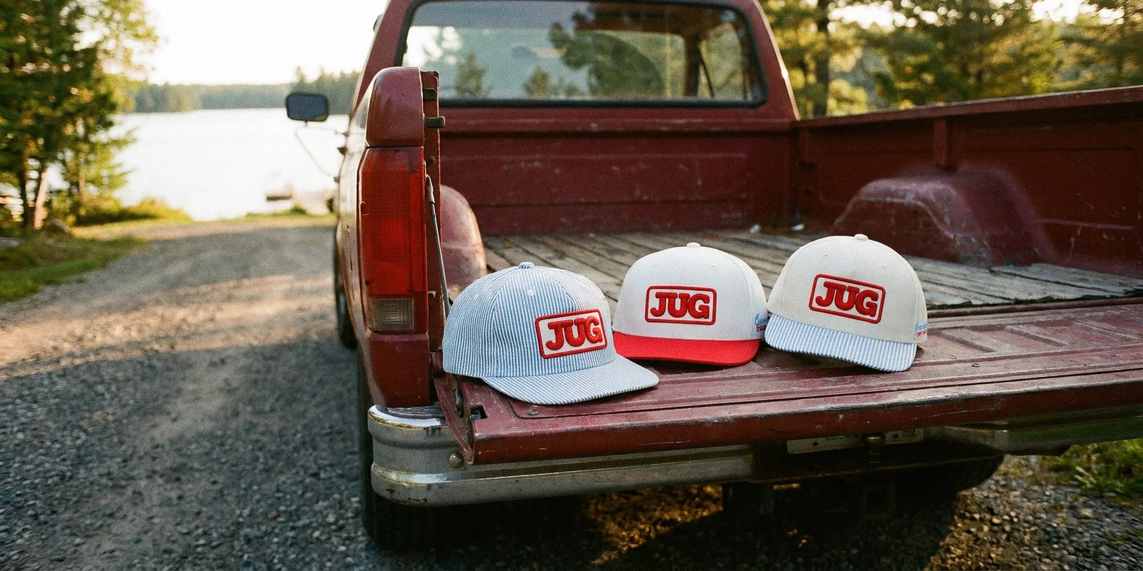 Three JUG canned water custom 5-panel hats lined up on the tailgate of a red pickup truck by a lake