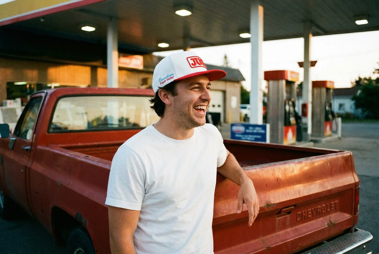 A man in a JUG Red Top Hat leaning on a red pickup truck at a gas station