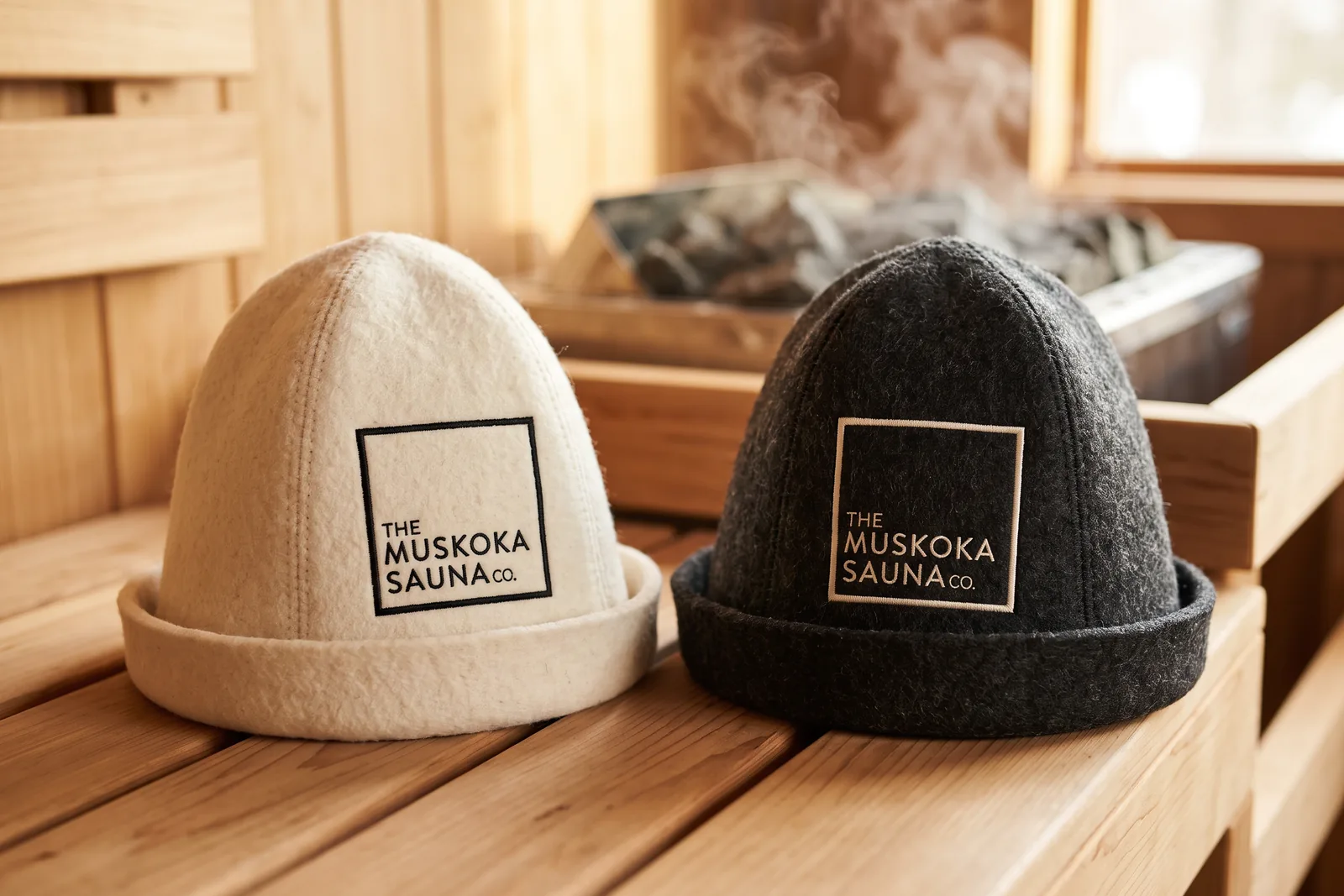 Two wool felt sauna hats, off-white and charcoal, with The Muskoka Sauna Co square logo embroidered on the front, on a cedar sauna bench