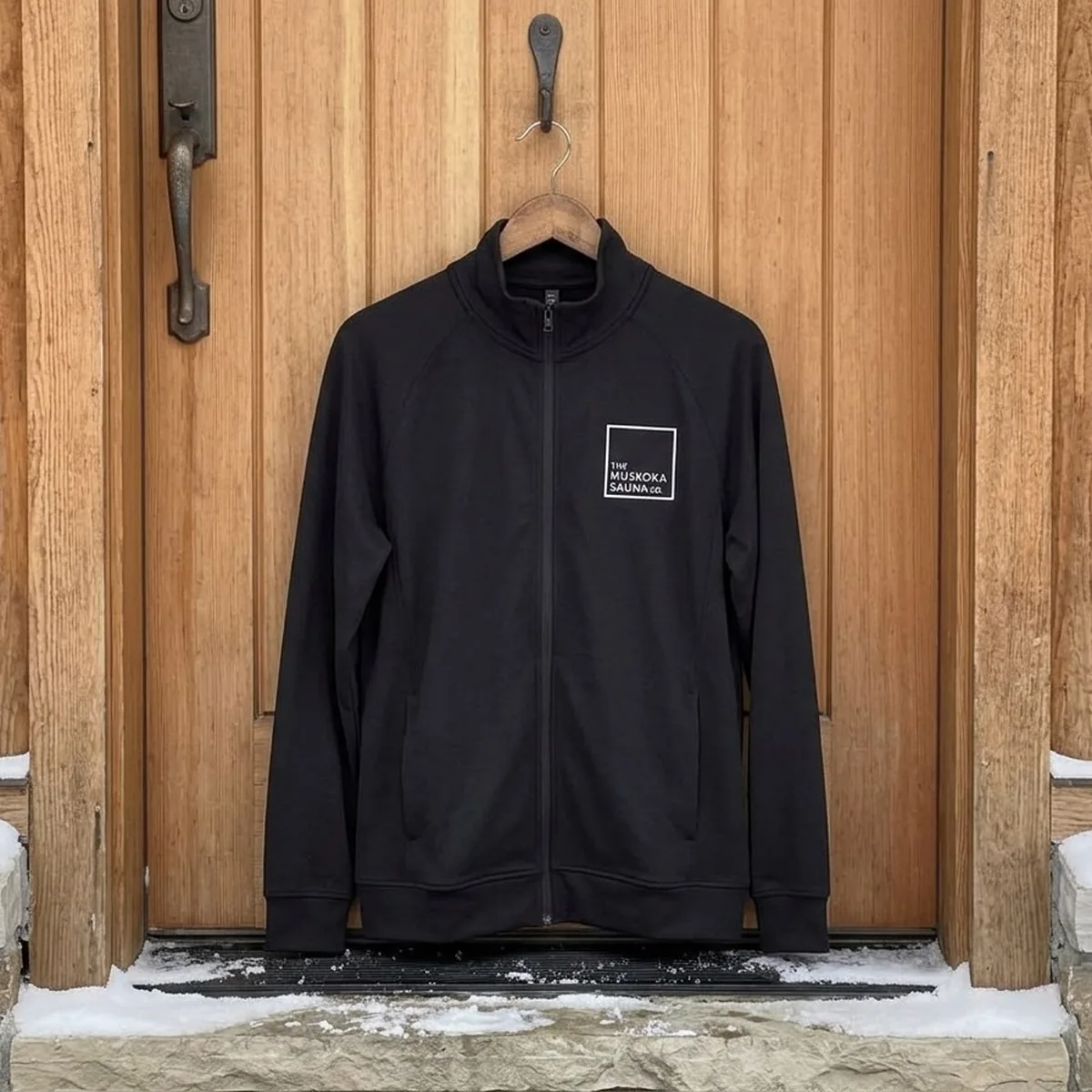 The Muskoka Sauna Co black jacket hanging on a cedar door in winter