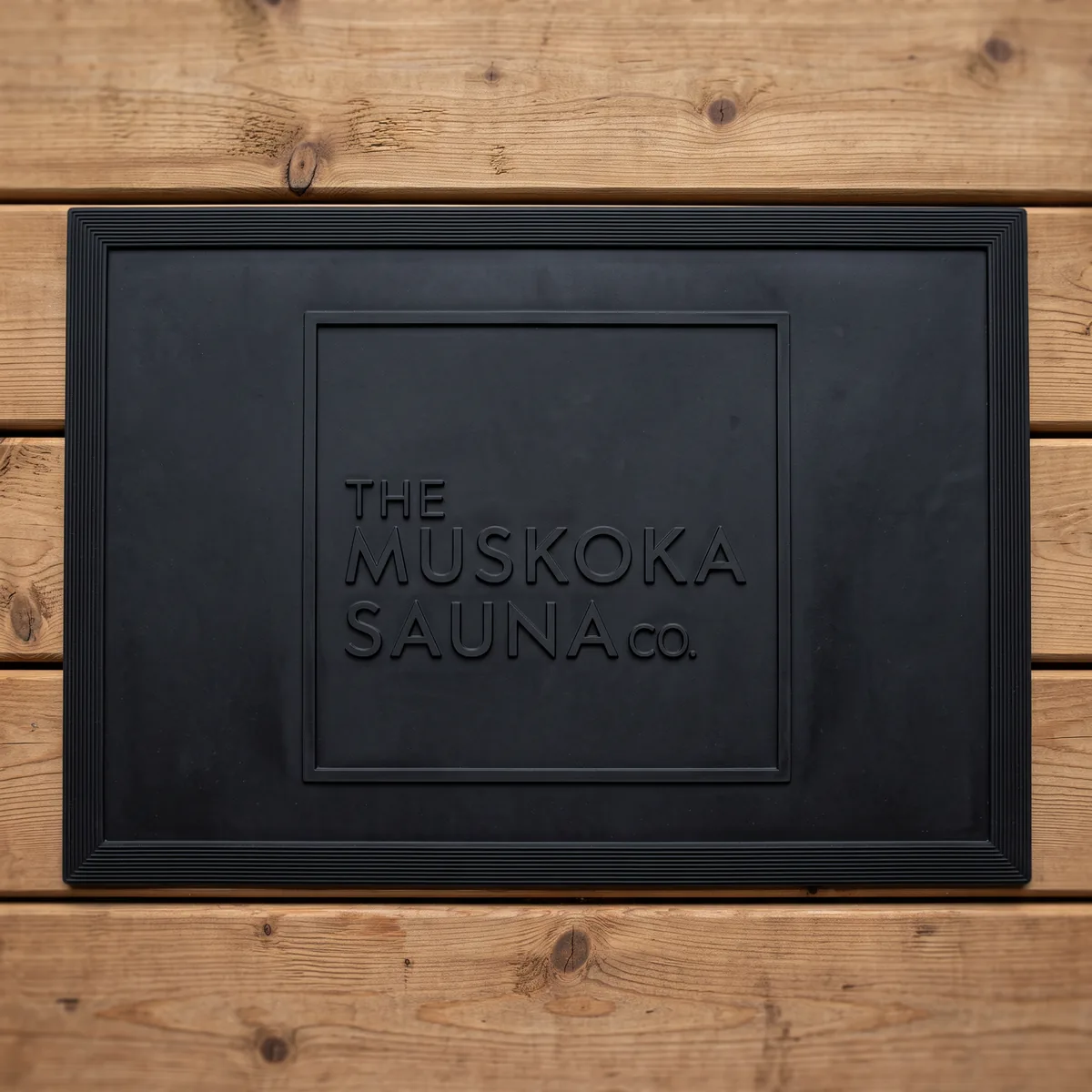 Black outdoor rubber sauna mat with The Muskoka Sauna Co square logo raised in the rubber, on cedar decking